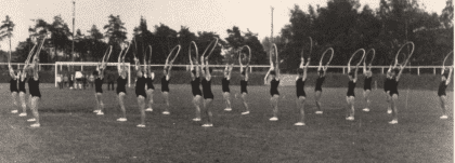 Tanzgruppe in Schwarz, die synchron mit Reifen auf einem Rasenfeld &uuml;bt, umgeben von B&auml;umen und Zuschauern.