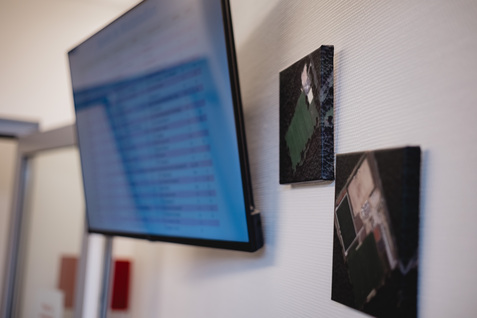 Flachbildschirm an der Wand mit Tabellenansicht, daneben zwei Bilder von Gr&uuml;nfl&auml;chen.