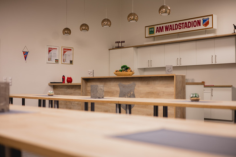 Moderner Seminarraum mit Holztheke, Dekoration und fest installierten Leuchten, im Hintergrund ein Schild mit "AM WALDSTADION".