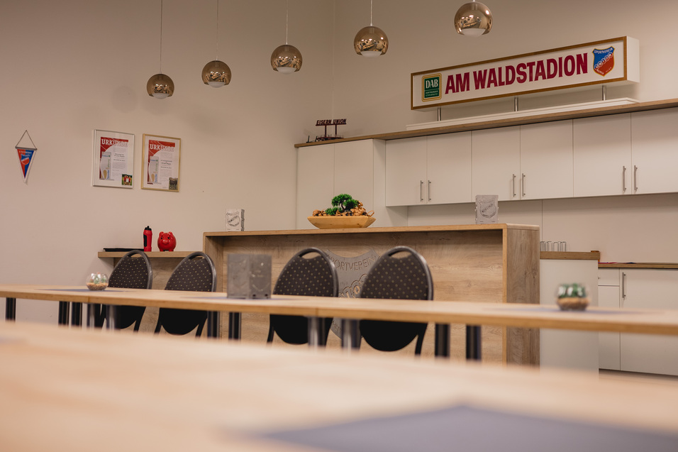 Modernes Besprechungszimmer mit Holz-Tisch, St&uuml;hlen, K&uuml;che und Wandschildern im Hintergrund.
