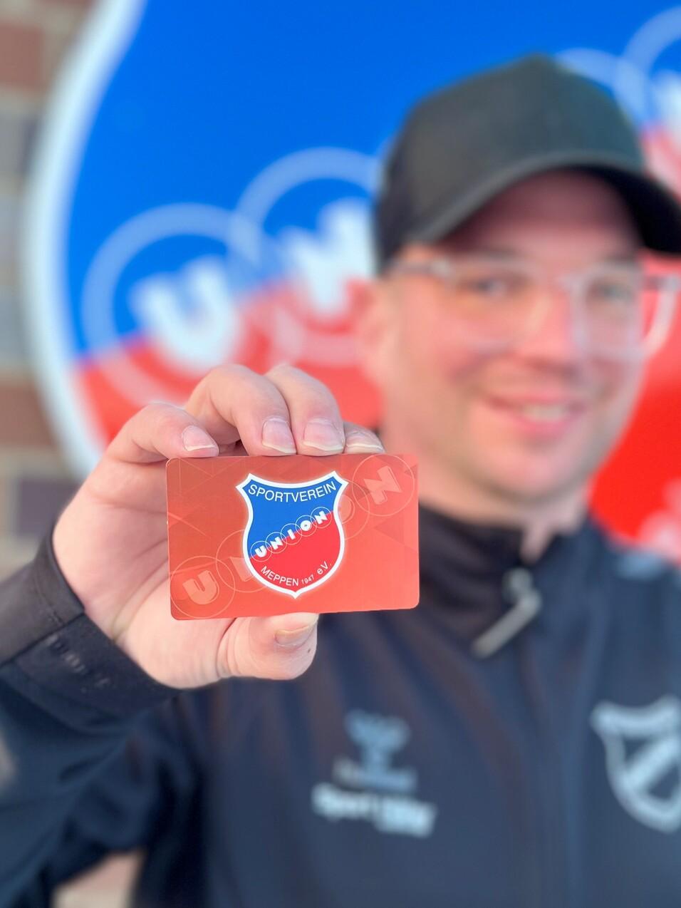 Mann zeigt eine Mitgliedskarte vor einem Sportverein-Logo an einer gemauerten Wand.
