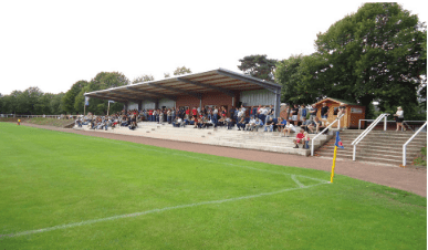 Zuschauer sitzen auf einer Trib&uuml;ne neben einem Fu&szlig;ballplatz, umgeben von B&auml;umen und einem Sportgeb&auml;ude.