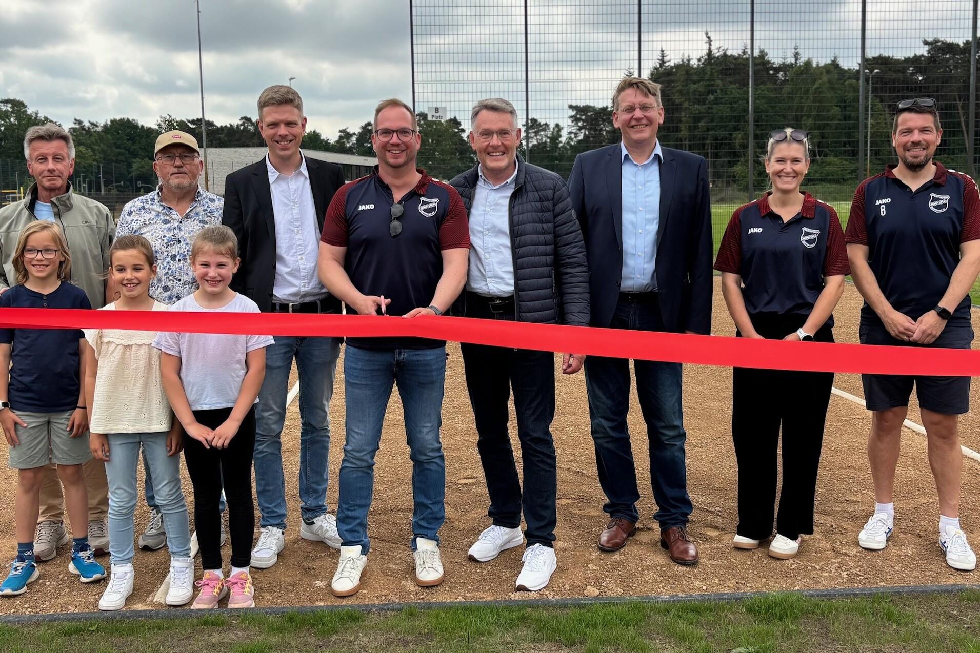 Er&ouml;ffnungszeremonie mit Personen und Roten Band auf einem Sportplatz, umgeben von Kindern und Erwachsenen.