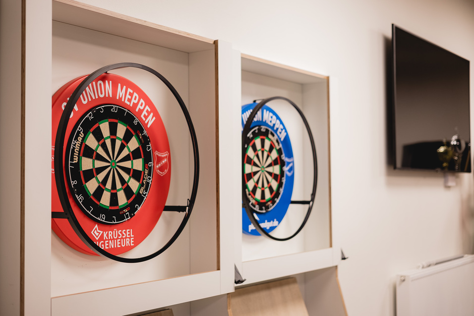 Zwei Dartscheiben in Rot und Blau, stilvoll in einer wei&szlig;en Wandnische montiert, mit einem TV im Hintergrund.