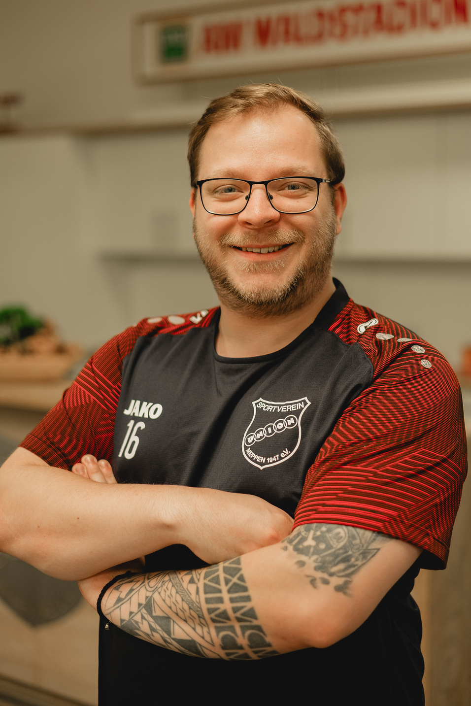 L&auml;chelnder Mann mit Brille, in sportlichem Trikot mit Vereinslogo, steht mit verschr&auml;nkten Armen vor einer K&uuml;chenzeile.