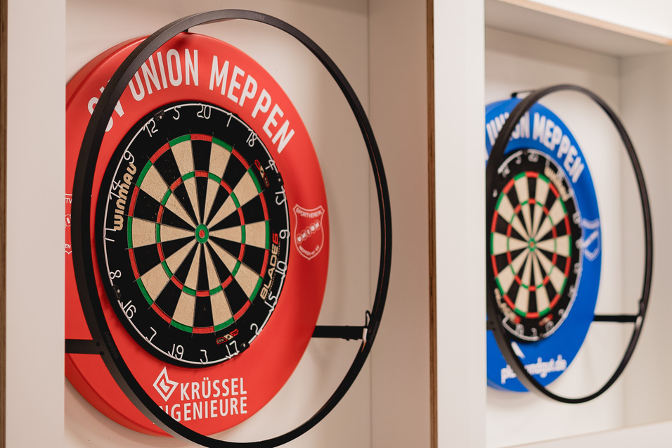 Zwei Dartscheiben in Rot und Blau, stilvoll in einer wei&szlig;en Wandnische montiert, mit einem TV im Hintergrund.