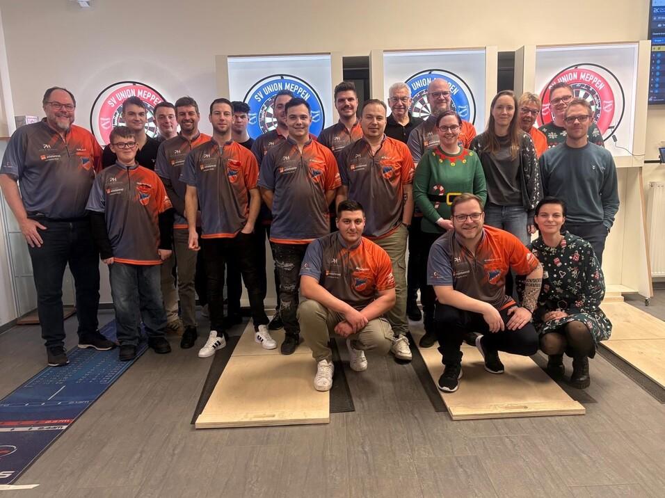 Gruppe von 16 Personen in Sportbekleidung posiert vor zwei gro&szlig;en Zielscheiben in einem Innenraum.