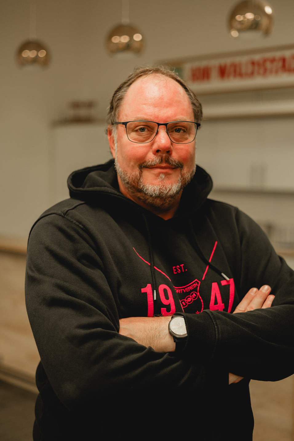 Mann mit Brille und Bart in schwarzem Hoodie steht mit verschr&auml;nkten Armen vor einer modernen K&uuml;che.