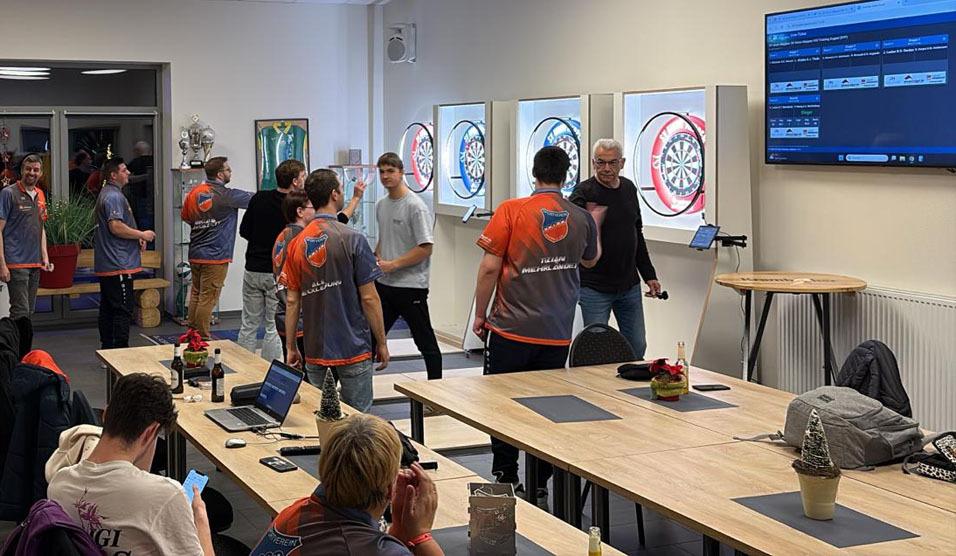 Gruppe von Dartspielern in einem Raum mit Dartboards und einem Bildschirm, Tische und Getr&auml;nke im Vordergrund.