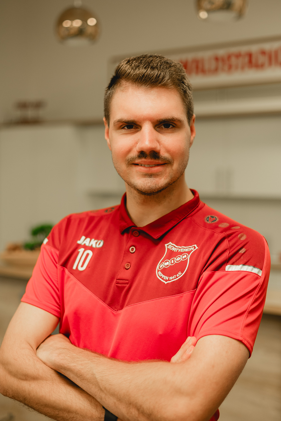 Portr&auml;t eines Mannes in rotem Sportpoloshirt mit Nummer 10, verschr&auml;nkt die Arme und l&auml;chelt selbstbewusst.