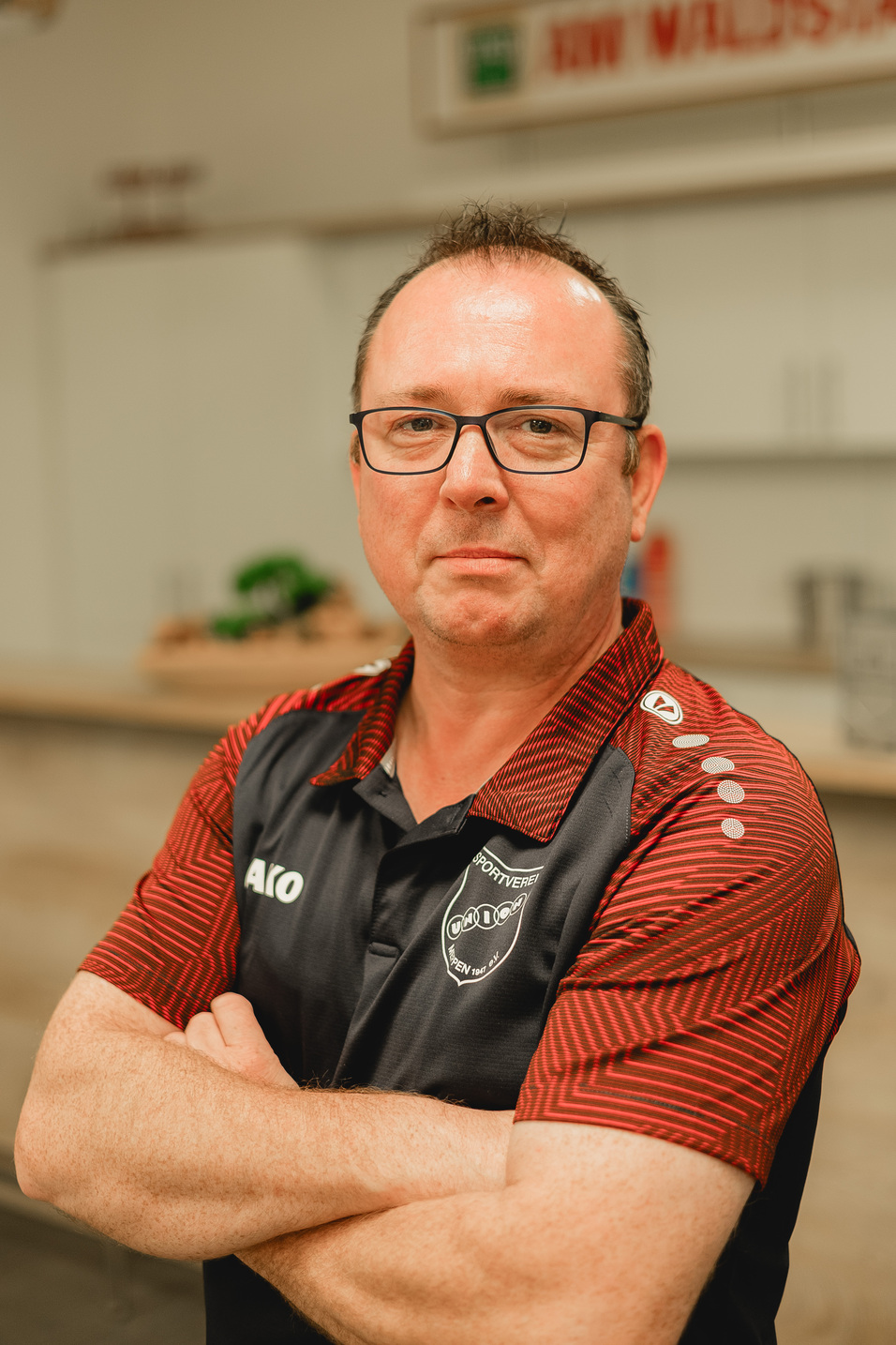 Mann mit Brille und gefalteten Armen in einem schwarz-roten Poloshirt steht vor einer hellen K&uuml;che.