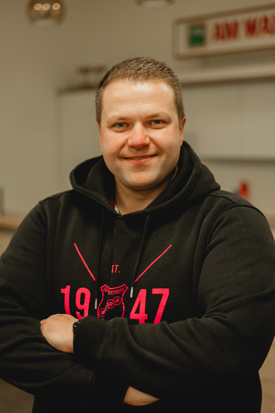 Portr&auml;t eines Mannes mit kurzen Haaren, der in einem schwarzen Hoodie mit rotem Druck l&auml;chelt und die Arme gekreuzt.