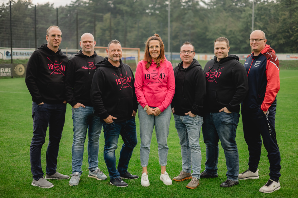 Gruppe von sieben Personen auf einem Sportplatz, drei M&auml;nner tragen dunkle Sweatshirts, eine Frau in pinkem Hoodie steht in der Mitte.