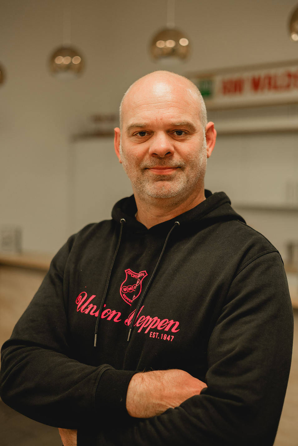 Mann mit kurzen grauen Haaren tr&auml;gt schwarzen Kapuzenpullover mit pinkem Logo, steht in moderner Innenraum.