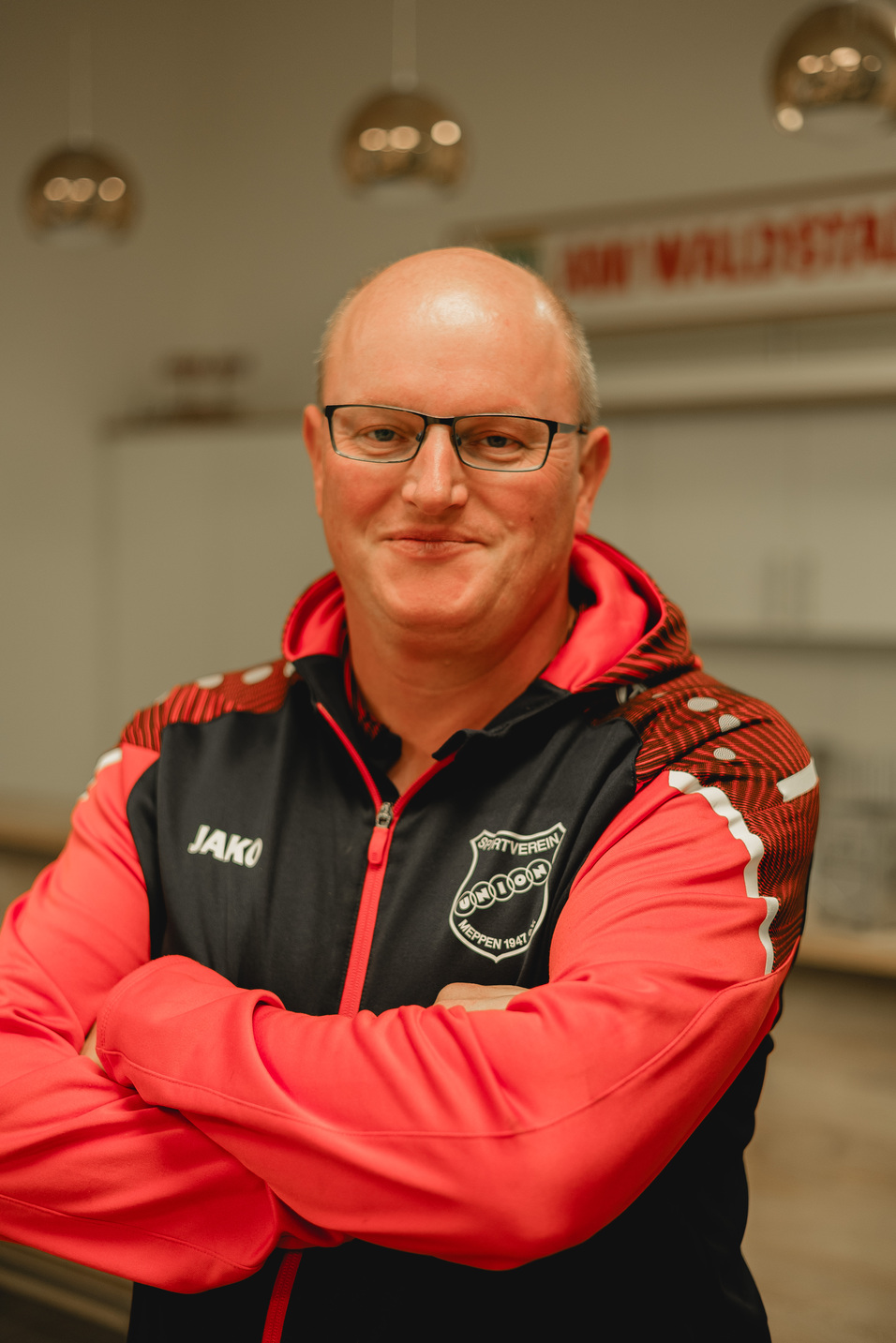 Mann mit Brille und kurzen Haaren in rot-schwarzer Sportjacke, l&auml;chelt mit verschr&auml;nkten Armen vor modernem Hintergrund.