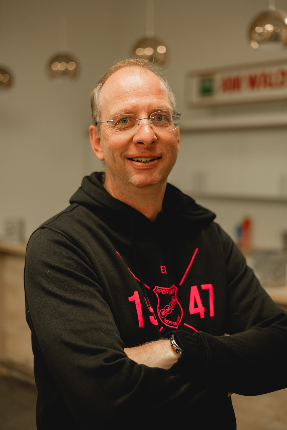 L&auml;chelnder Mann mit Brille und Hoodie steht leicht schr&auml;g, Arme vor der Brust verschr&auml;nkt, in hellem Innenraum.