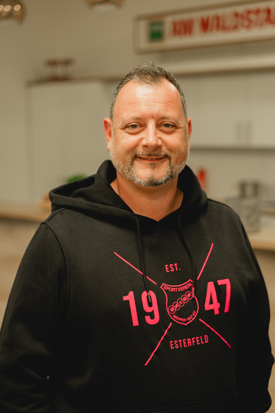 Mann mit Bart und schwarzem Kapuzenpullover steht vor einer hellen Innenraumatmosph&auml;re.