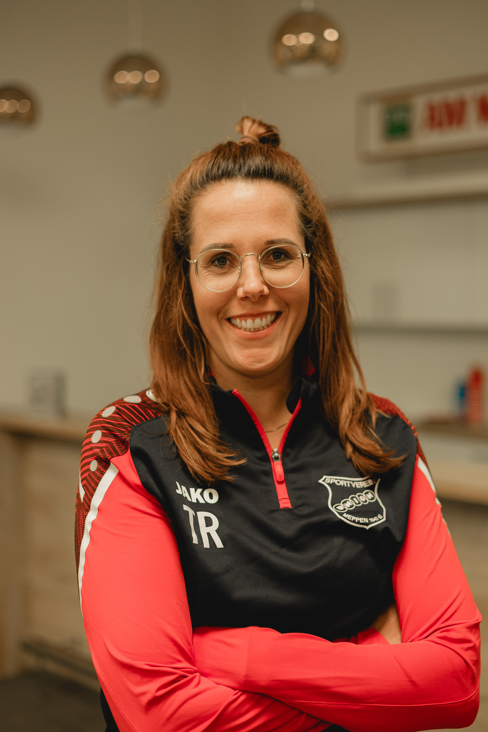 Portr&auml;t einer l&auml;chelnden Frau mit langen, braunen Haaren, die ein sportliches Oberteil mit roten und schwarzen Farben tr&auml;gt.