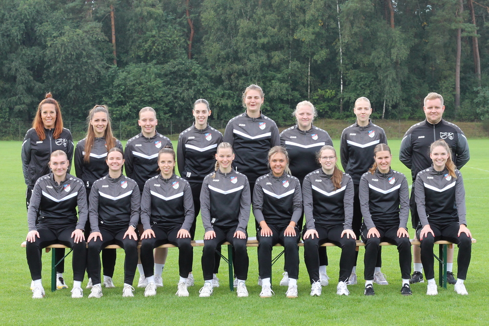 Mannschaftsbild einer Frauenfu&szlig;ballmannschaft in schwarzen Trainingsanz&uuml;gen, auf einer gr&uuml;nen Wiese posierend.