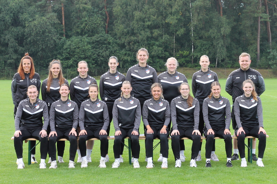 Mannschaftsbild einer Frauenfu&szlig;ballmannschaft in schwarzen Trainingsanz&uuml;gen, auf einer gr&uuml;nen Wiese posierend.