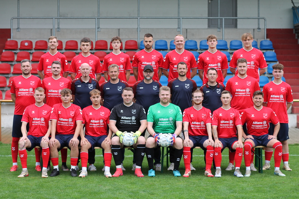 Mannschaftsbild einer Fu&szlig;ballmannschaft in roten Trikots, auf einem Sportplatz mit Trib&uuml;ne im Hintergrund.