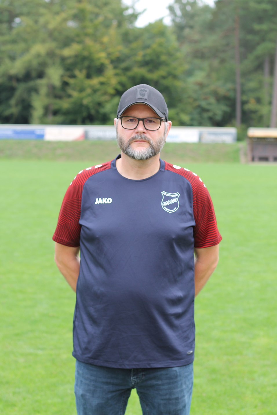 Mann mit Brille und grauem Bart steht in einem Sporttrikot auf einem gr&uuml;nen Fu&szlig;ballplatz.