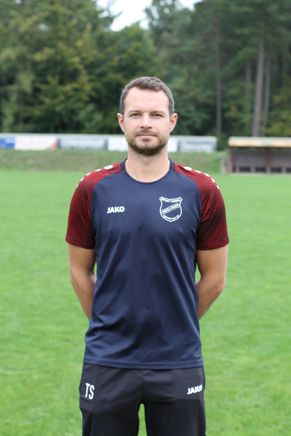 Portr&auml;t eines Mannes in Sportbekleidung, stehend auf einem Fu&szlig;ballplatz mit gr&uuml;ner Wiese im Hintergrund.