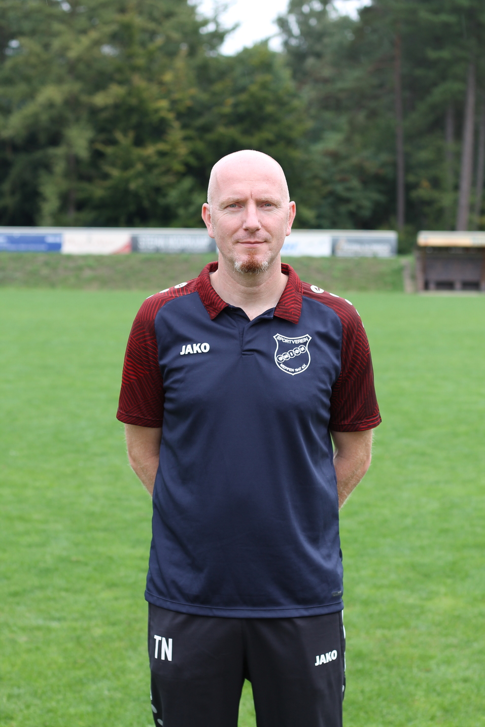 Mann mit Glatze steht auf einem Fu&szlig;ballplatz, tr&auml;gt ein poloshirt mit Vereinslogo und Shorts. Hintergrund ist bewaldet.