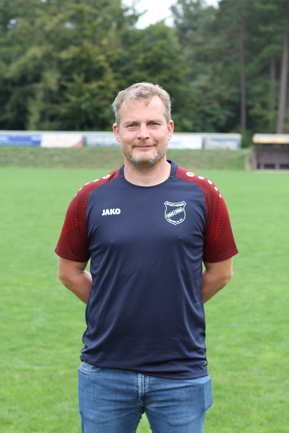 Mann mit sportlichem Oberteil steht auf einem Fu&szlig;ballplatz mit gr&uuml;ner Wiese und B&auml;umen im Hintergrund.