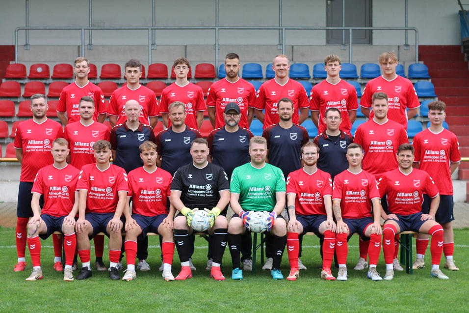 Mannschaftsbild einer Fu&szlig;ballmannschaft in roten Trikots, auf einem Sportplatz mit Trib&uuml;ne im Hintergrund.