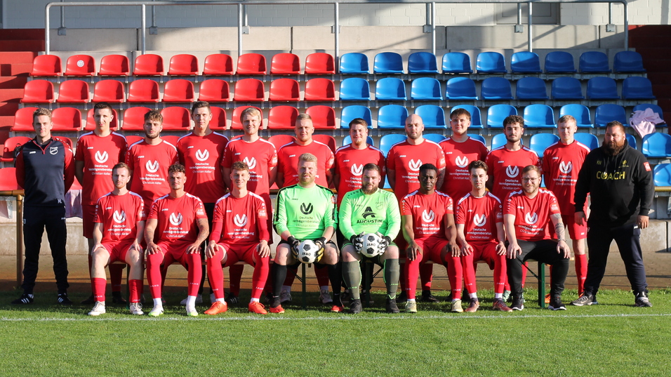 Teamfoto einer Fu&szlig;ballmannschaft in roten Trikots, sitzend und stehend auf einer Stadiontrib&uuml;ne mit blauen und roten Sitzen.