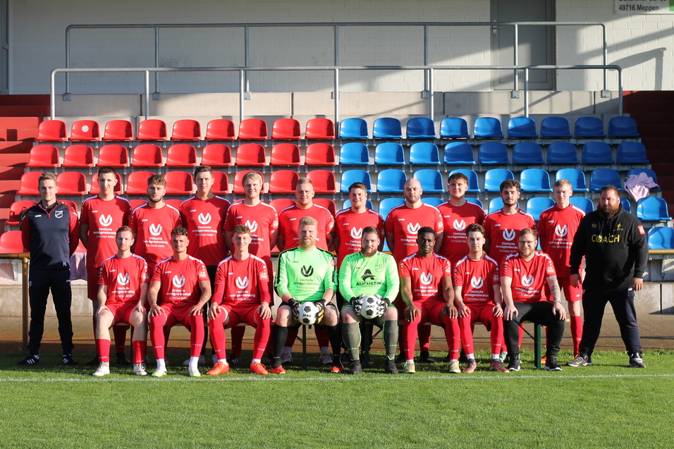 Teamfoto einer Fu&szlig;ballmannschaft in roten Trikots, sitzend und stehend auf einer Stadiontrib&uuml;ne mit blauen und roten Sitzen.