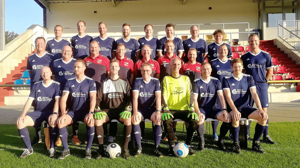 Mannschaftsfoto einer Fu&szlig;ballmannschaft in blauen Trikots auf einem Sportplatz, mit Torwart in gelbem Trikot.