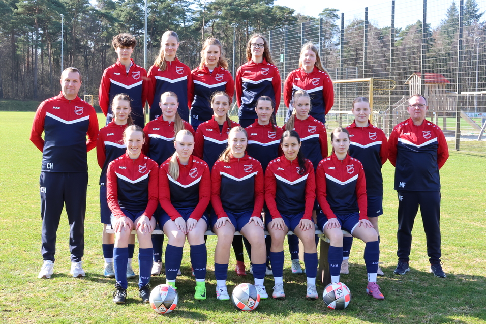 Gruppenfoto einer Fu&szlig;ballmannschaft mit 15 Spielerinnen in roten und blauen Trikots auf einem gr&uuml;nen Sportplatz.