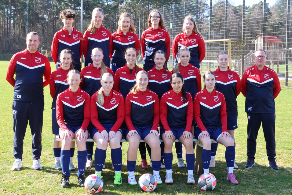 Gruppenfoto einer Fu&szlig;ballmannschaft mit 15 Spielerinnen in roten und blauen Trikots auf einem gr&uuml;nen Sportplatz.
