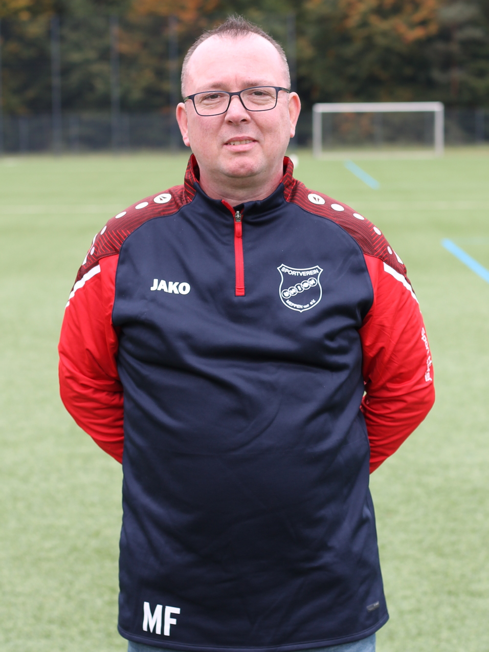 Mann im Sportoutfit mit Brille steht auf einem Fu&szlig;ballplatz, im Hintergrund ein Tor und bunte B&auml;ume.