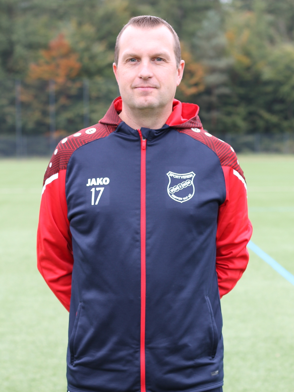 Portr&auml;t eines Mannes in Sportkleidung mit rot-schwarzer Jacke auf einem Fu&szlig;ballfeld im Hintergrund.
