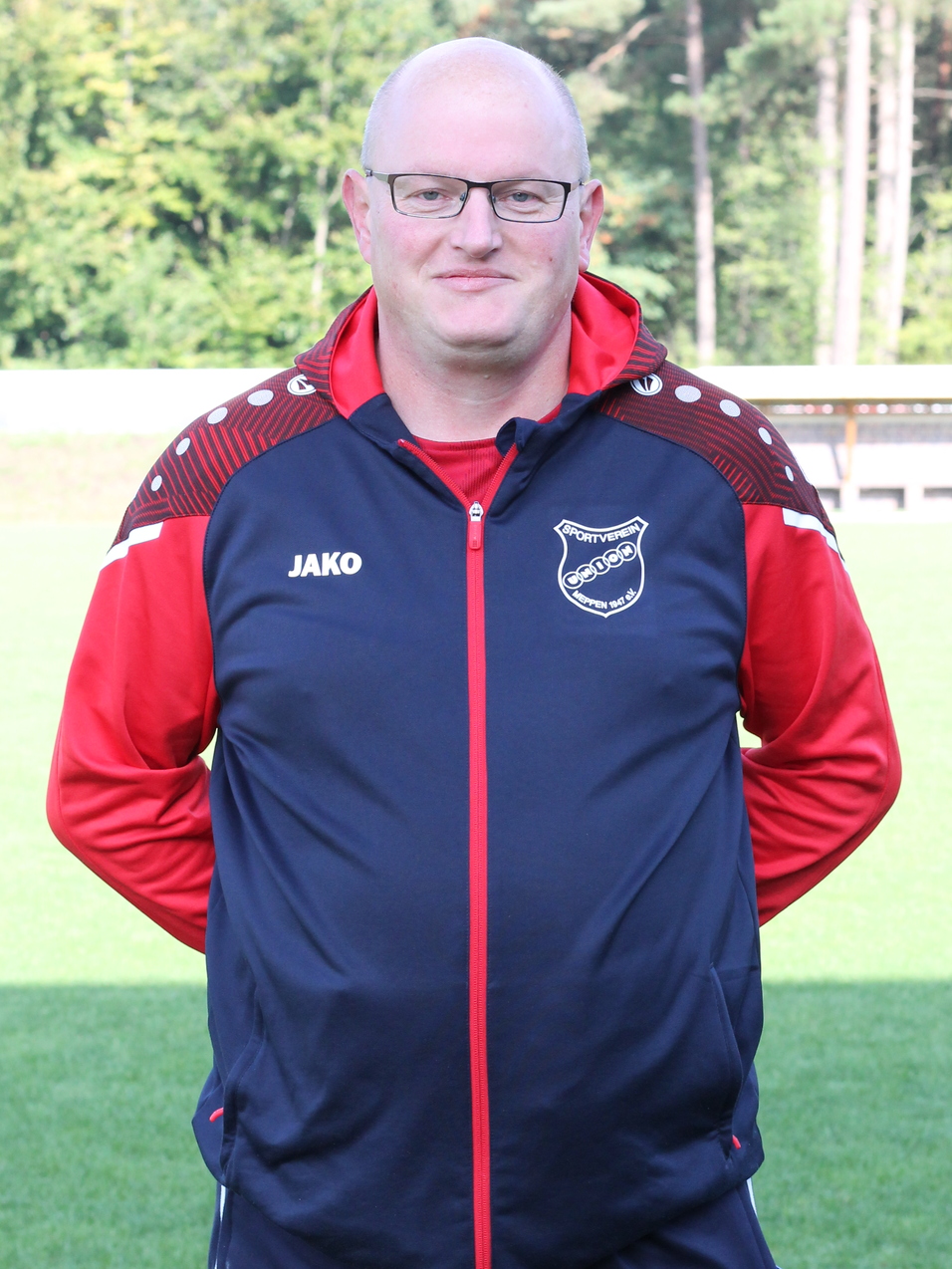 Trainer in Sportanzug mit Brille, steht auf einem gr&uuml;nen Fu&szlig;ballplatz vor einem verschwommenen Hintergrund.