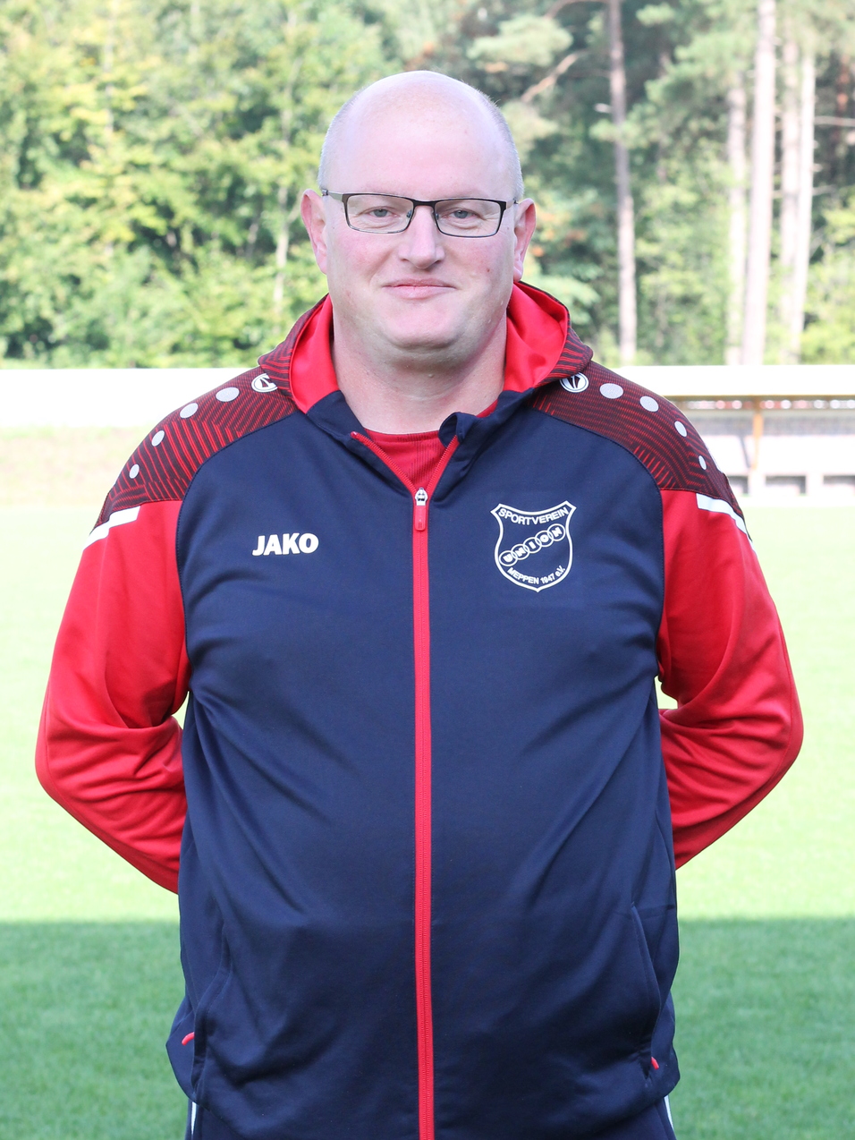 Trainer in Sportanzug mit Brille, steht auf einem gr&uuml;nen Fu&szlig;ballplatz vor einem verschwommenen Hintergrund.