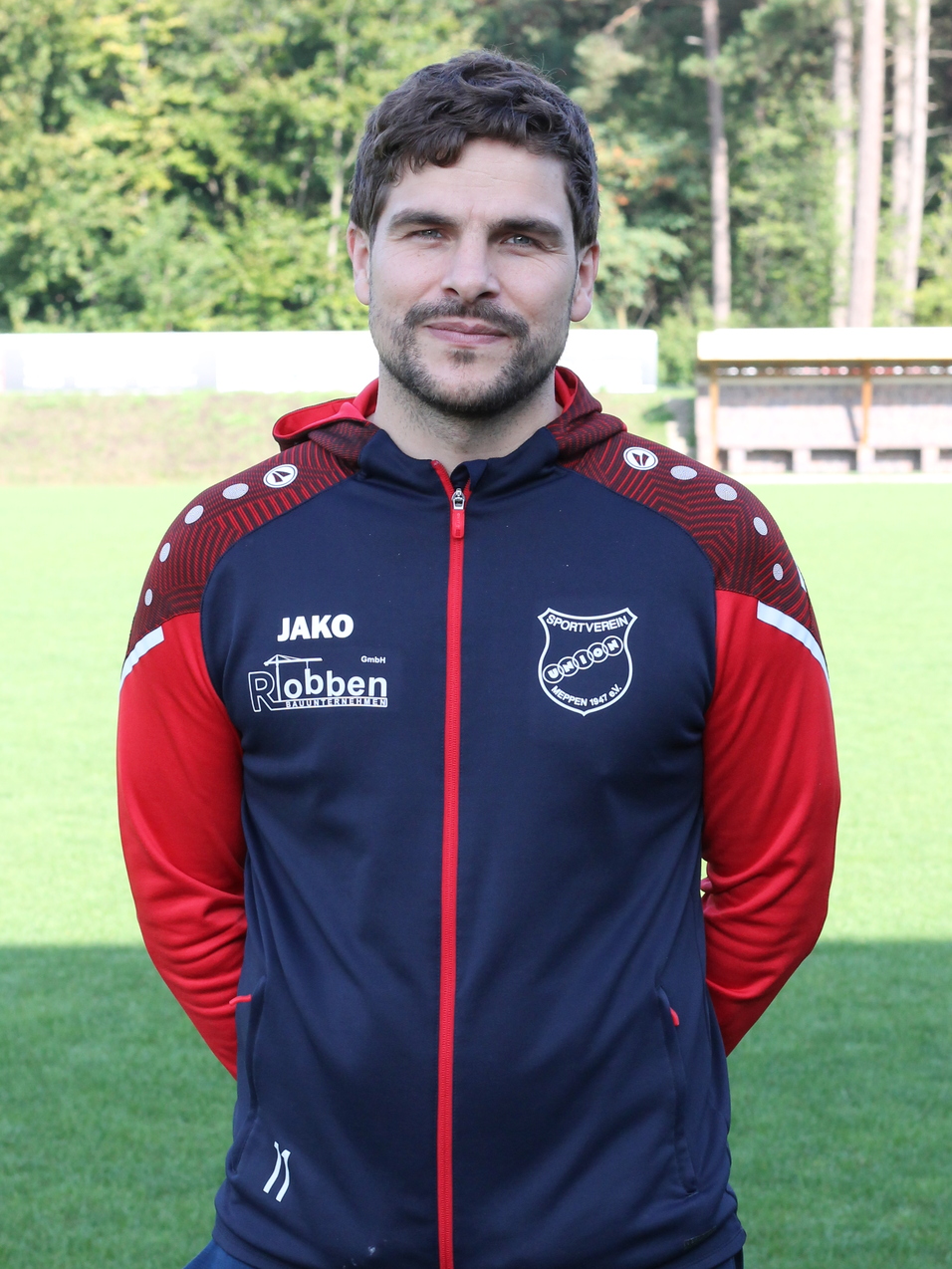 Mann in Sportjacke steht auf einem Fu&szlig;ballplatz, im Hintergrund B&auml;ume und eine Trib&uuml;ne.