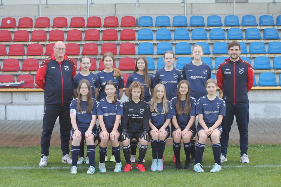 Mannschaftsfoto einer Fu&szlig;balljugend mit Trainer, in schwarzen Trikots, vor einer Trib&uuml;ne mit roten und blauen Sitzen.