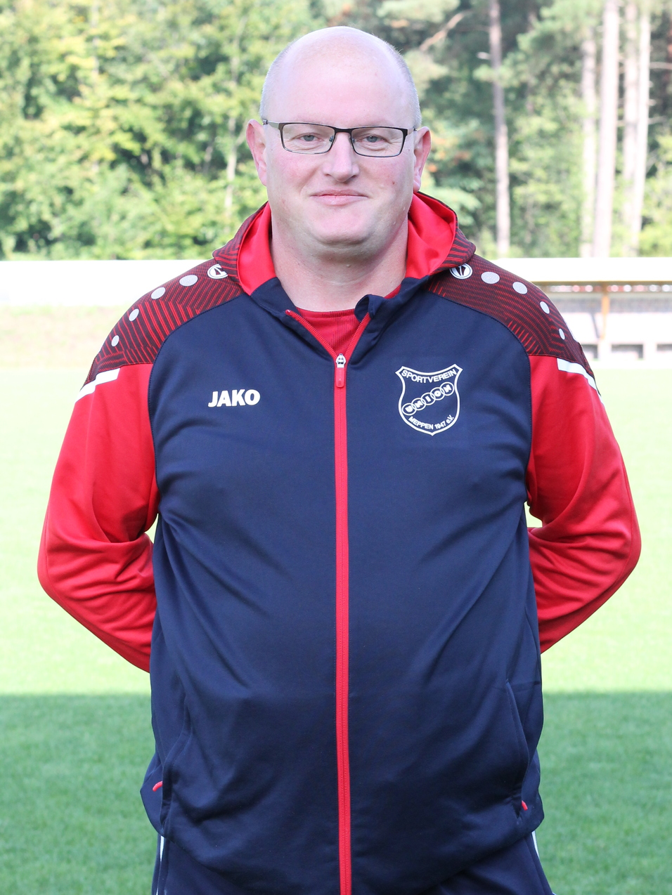 Trainer in Sportanzug mit Brille, steht auf einem gr&uuml;nen Fu&szlig;ballplatz vor einem verschwommenen Hintergrund.