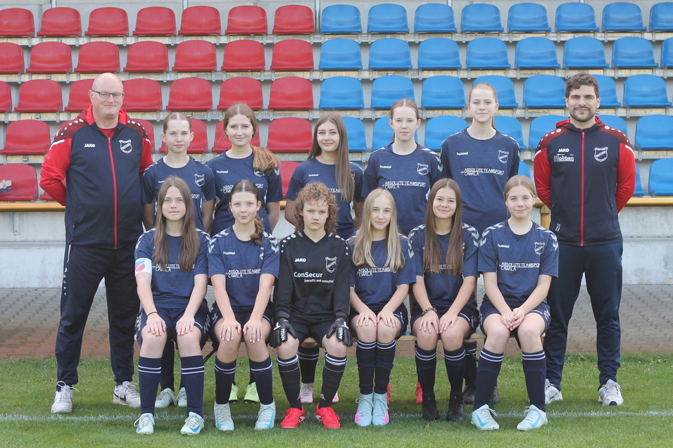 Mannschaftsfoto einer Fu&szlig;balljugend mit Trainer, in schwarzen Trikots, vor einer Trib&uuml;ne mit roten und blauen Sitzen.