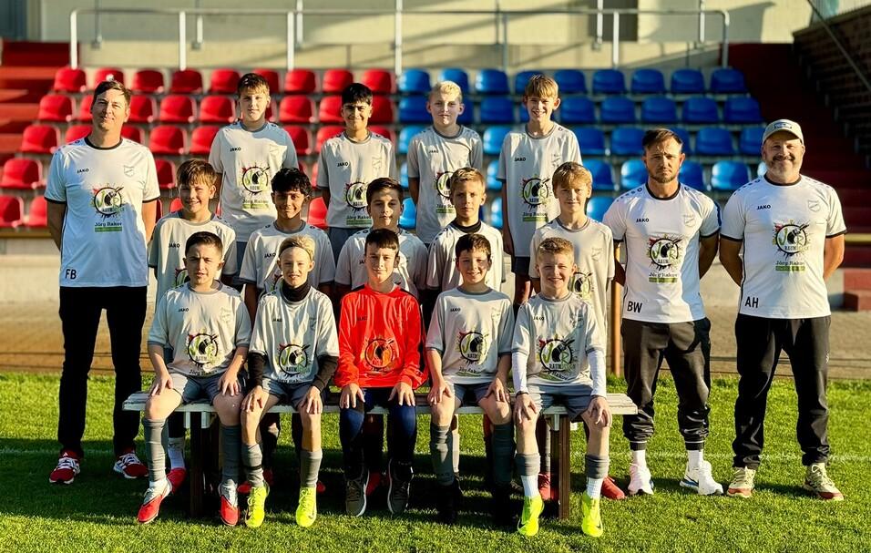 Portr&auml;t einer Fu&szlig;ballmannschaft aus 15 Spielern, ein Torwart in rotem Trikot, auf einem Sportplatz vor Trib&uuml;nen.