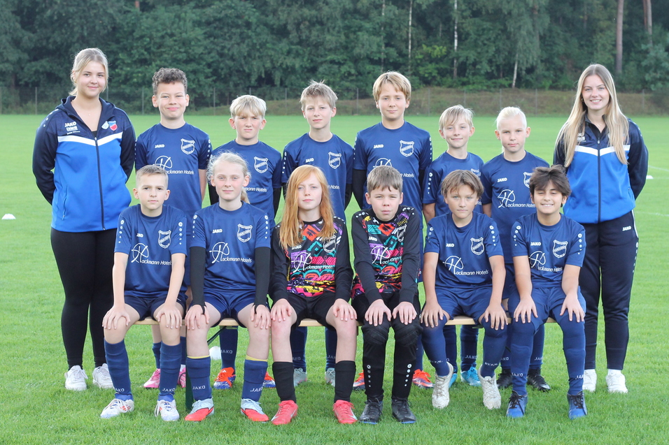 Gruppenfoto einer Fu&szlig;ballmannschaft mit Trainerinnen auf einem gr&uuml;nen Rasenplatz, alle in blauen Trikots.