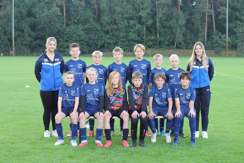 Gruppenfoto einer Fu&szlig;ballmannschaft mit Trainerinnen auf einem gr&uuml;nen Rasenplatz, alle in blauen Trikots.