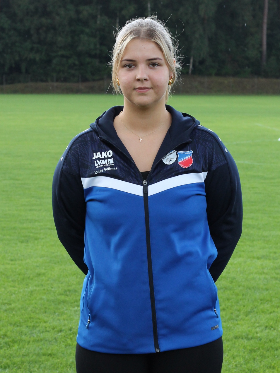 Junge Frau in blauer Sportjacke steht auf einem gr&uuml;nen Fu&szlig;ballfeld mit B&auml;umen im Hintergrund.
