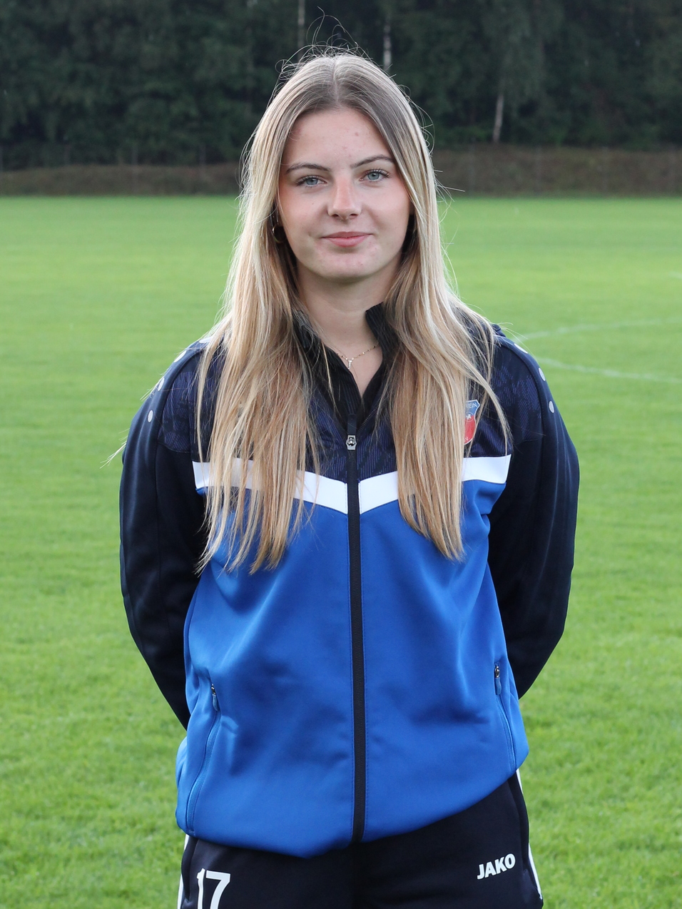 Junge Frau mit langen Haaren steht auf einem gr&uuml;nen Sportfeld, tr&auml;gt Trainingsanzug in Blau und Schwarz.