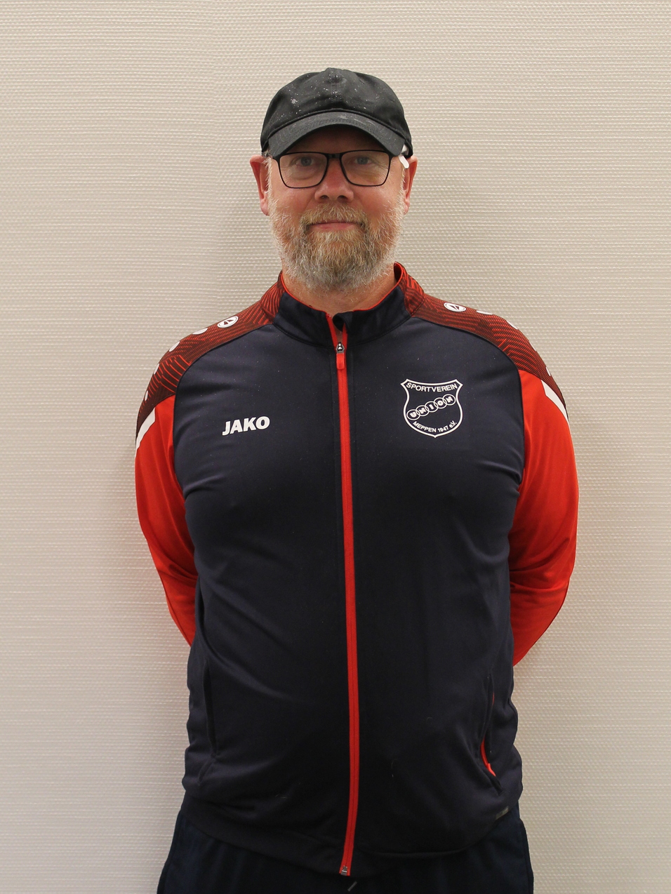 Mann mit Brille und Bart in Sportjacke mit Logo steht vor einer hellen Wand.