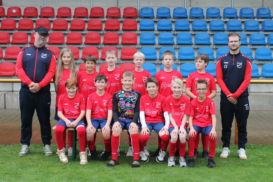 Jugendfu&szlig;ballmannschaft in roten Trikots steht auf dem Rasen, betreut von zwei Trainern im Hintergrund.